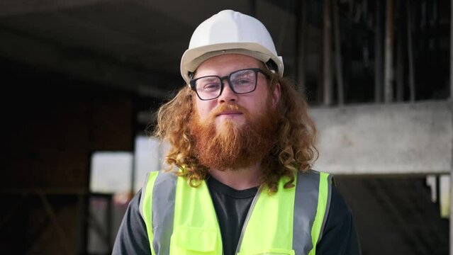 Close up face construction worker with smile in uniform and helmet smiling looking at camera, bearded man engineer architect wearing protective hardhat and vest at construction site. 