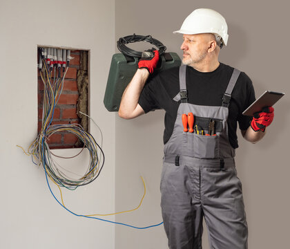 Man electrician. Guy holding coil of wires. Electrical engineer near place for power panel. Editor with tablet and tool case. Man wants to install electrical panel. Electrician in gray overalls