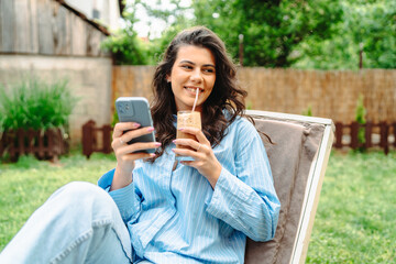 One young caucasian girl is relaxing on easy chair while drinking coffee and using mobile phone in...
