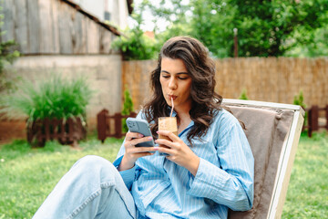 One young caucasian girl is relaxing on easy chair while drinking coffee and using mobile phone in...
