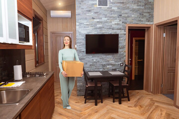 Woman with box enters house. Girl holding cardboard parcel. Moving box in hands of lady. Housewarming process. Female moving into new home. Woman with box in apartment kitchen. Change housing.