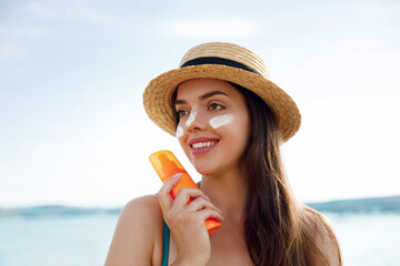 Beautiful young woman at beach applying sunscreen on face and looking at camera. Beauty girl...