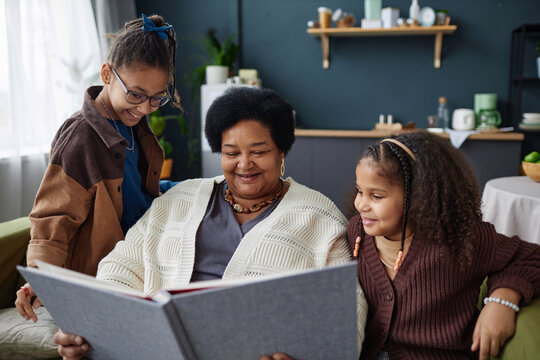 Portrait of smiling senior African American woman with two children looking at family photo album together at home - Powered by Adobe