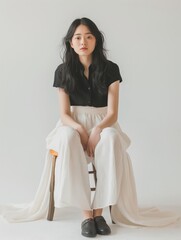 A serene young Asian woman is seated on a simple wooden stool in a bright room with a white background.