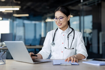 A professional Asian female doctor is smiling while using a laptop in a bright, contemporary office setting. She is equipped with a stethoscope and medical attire, appearing competent and approachable