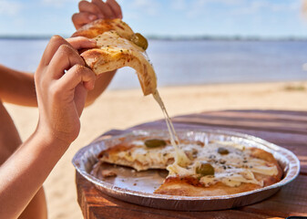 child's hands holding a slice of mozzarella pizza, with the sand and river in the background