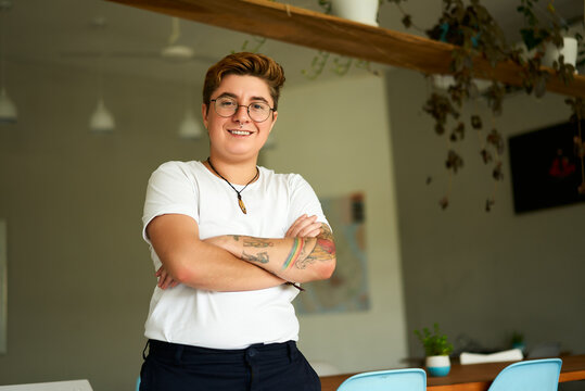 Confident Gen Z transgender person stands in modern office, arms crossed, smiling. Inclusive workplace with natural light, plants. Casual work attire, LGBTQ pride, authenticity at work reflected.