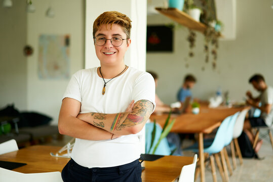 Confident transgender male employee smiling in office space, arms crossed, diverse team working in background. Inclusion at modern workplace, LGBTQ representative