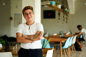 Confident transgender pro in casual office attire stands with arms crossed. Warm, welcoming smile...
