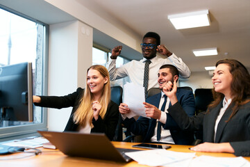Multiethnic business team in office, jubilant at computer screen success. Cheerful diverse colleagues with laptop enjoy corporate victory. Excited mixed group, male, female, celebrate project win.