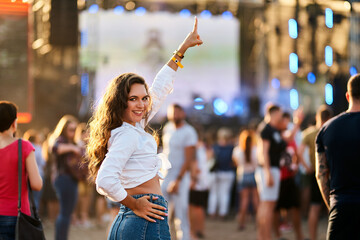 Smiling young woman enjoys live music at beach festival, waving hand, dancing among crowd. Summer...