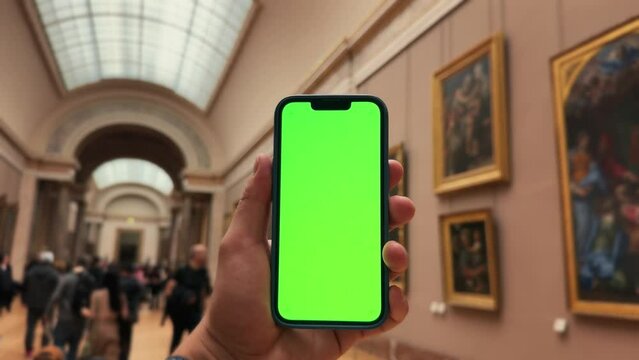 Close up of woman's hand holds phone with green screen mock up. Blurred masterpiece art at background. Concept of digital app for museums and virtual excursion.
