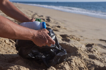 Close-up of a man's tanned hands picking up garbage in a black bag on the shore of a beautiful...