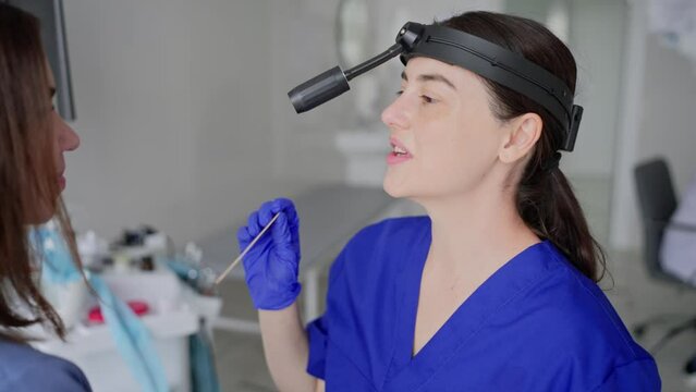 Over the shoulder of a confident brunette girl. An ENT doctor in a blue uniform puts a wooden stick in a girls mouth to check for throat diseases in a modern clinic
