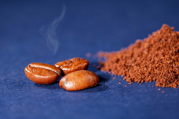 Three coffee beans with steam and a pile of ground coffee on a blue background