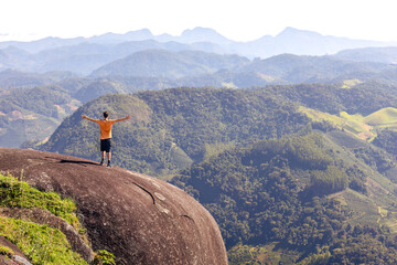 Man with open arms on edge of a cliff, radiating well-being and joy as he takes in the mountain landscape. Reflects pursuit of personal growth and well-being through travel and adventurous experiences