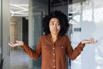 Young African American female professional in a polka dot blouse, expressing confusion and uncertainty. Indoor office setting with natural light reflecting off glass background.