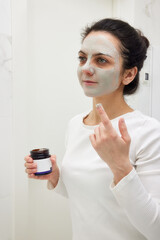Caucasian woman looking in mirror and applying face mask white in bathroom