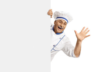 Mature male chef in a uniform waving from behind a blank panel