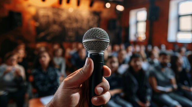 Close-up, hand with microphone ready to speak, blurred attendees of a classroom in the background, studio lighting isolates the subject