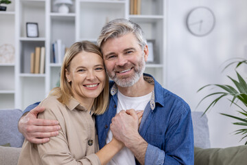 A cheerful middle-aged couple hugs warmly in their stylish living room, smiling at the camera with a bookshelf and modern decor in the background.