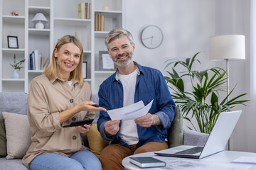 A mature couple sitting on a couch, smiling as they discuss documents and financial plans together,...