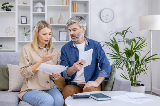 A mature couple sits closely on a sofa in a well-appointed living room, discussing documents with concern and concentration.