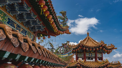 Details of the architecture of a beautiful Chinese temple. The tiled curved roofs are decorated with ornaments against a blue sky, clouds. Rows of traditional red lanterns are suspended along the edge
