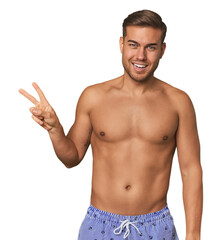 Fototapeta premium Young man shirtless in swimwear, studio joyful and carefree showing a peace symbol with fingers.