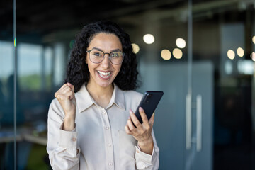 Cheerful young businesswoman in eyeglasses celebrates a victory with her smartphone in an office. She is showing a joyful expression and fist pump.