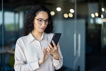 A focused Hispanic businesswoman in glasses uses her smartphone, possibly reading an important...