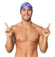Young Hispanic man with swim gear indicates with both fore fingers up showing a blank space.