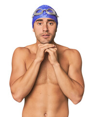 Young Hispanic man with swim gear praying for luck, amazed and opening mouth looking to front.