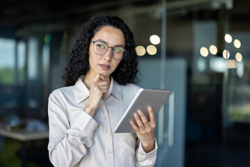A professional Hispanic businesswoman thinking deeply while holding a digital tablet in a modern...