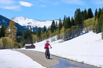 smiling senior woman riding her electric mountain bike on a sunny day in early spring in the Gunzesried Valley below the snow capped high Allgaeu mountains in background
 