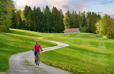 pretty senior woman riding her electric mountain bike in  springtime in the Allgau mountains near Oberstaufen,  with blooming yellow blooming Dandelion flowers in the Foreground, Bavaria, Germany