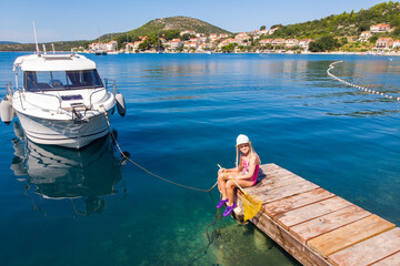 Cute little girl kid sits on wooden pier next moored boat yacht with scenic turquoise sea water...