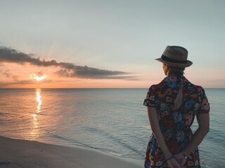Woman in hat and cute summer dress standing at the beach, looking at sunrise
