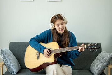 A woman is playing the guitar while wearing headphones. She is sitting on a couch. Concept of...