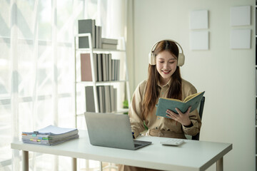 Fototapeta premium A woman is sitting at a desk with a laptop and a book. She is wearing headphones and smiling. The scene suggests that she is enjoying her work or leisure time