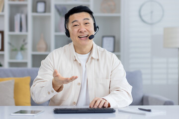 Cheerful Asian male in a beige shirt using a headset in a home office, looking at camera with a smile, possibly in a customer service call.
