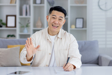 Cheerful Asian man sitting at home, talking and waving to the camera during an online meeting. He...