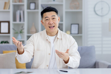 Cheerful Asian man engaging in a casual conversation at home, looking at the camera and talking with expressive hand gestures in a modern living room setting.