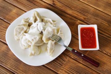 Cooked Russian dumplings in a white plate with a fork next to red tomato sauce on a table made of natural wooden pine boards. Photo