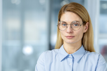 Confident professional businesswoman in a blue blouse and round glasses, standing in a modern...