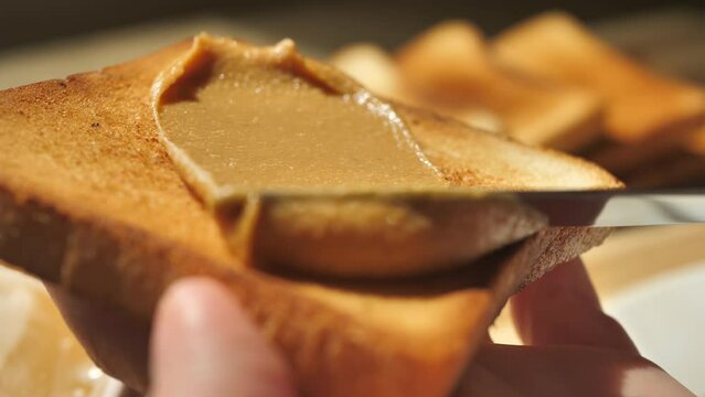 Spreading peanut butter onto crusty toasted bread using a table knife. Slow motion beautifully spread peanut butter on golden toast in the sunlight. Healthy breakfast with high-calorie foods