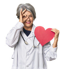 Caucasian mid-age doctor with heart symbol excited keeping ok gesture on eye.