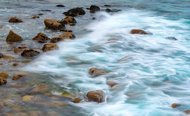 Long exposure of splashing breaking waves on the coast of California (USA). Shell-covered rocks and stones in the tidal current in the evening twilight at Lovers Point in Pacific Grove near Monterey.