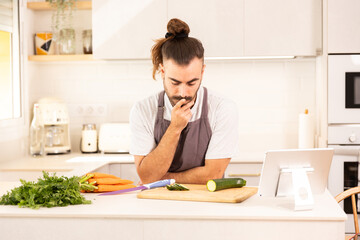 A man is sitting at a kitchen counter with a tablet in front of him. He is looking at a carrot and a cucumber on the cutting board