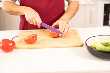 A man is cutting a tomato on a cutting board. The tomato is on the left side of the cutting board and the knife is on the right side